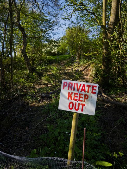 An outdoor woodland area with a narrow dirt path surrounded by green trees and foliage under a clear blue sky. In the foreground, a white rectangular sign with red text reading 'PRIVATE KEEP OUT' is mounted on a yellow wooden post, positioned beside a thin wire fence at the edge of the path. This scene suggests an access restriction or private property boundary. The sign and natural environment imply a context where house removals or movement services might need to navigate restricted or private grounds, as coordinated by companies like Man and Van Hampton for complex relocation or access-sensitive property removals.