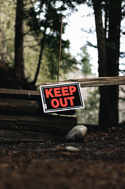 A small rectangular sign with a black background and bold red lettering reading 'KEEP OUT' is hanging from a thin string attached to a weathered wooden beam. The sign is positioned outdoors in a natural setting, with a backdrop of tall, blurred trees in green and brown tones. The ground beneath the sign is covered with dirt and small rocks, and a smooth, rounded stone is situated directly below the sign. This scene appears to depict a restricted access area, potentially related to property or land management, with natural elements suggesting a rural or wooded environment, aligning with house removals and property access solutions provided by Man and Van Hampton.