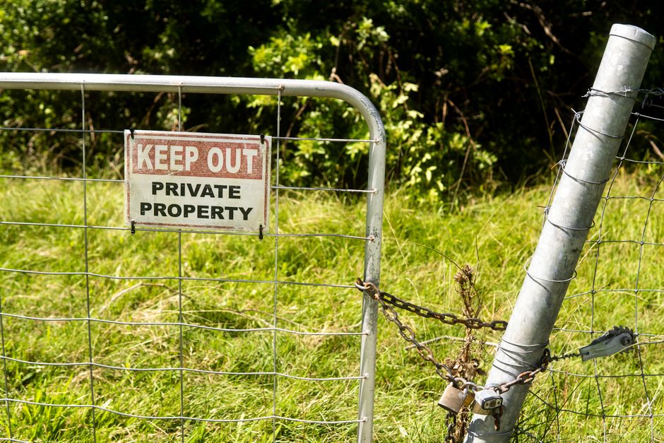 An outdoor woodland area with a narrow dirt path surrounded by green trees and foliage under a clear blue sky. In the foreground, a white rectangular sign with red text reading 'PRIVATE KEEP OUT' is mounted on a yellow wooden post, positioned beside a thin wire fence at the edge of the path. This scene suggests an access restriction or private property boundary. The sign and natural environment imply a context where house removals or movement services might need to navigate restricted or private grounds, as coordinated by companies like Man and Van Hampton for complex relocation or access-sensitive property removals.
