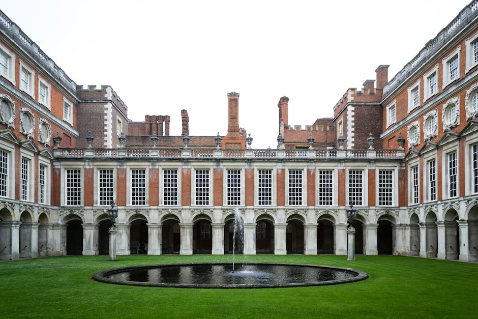 A view of the inner courtyard of a historic Georgian-style mansion, featuring a neatly maintained green lawn with a small, circular fountain at its center. Surrounding the courtyard is a symmetrical, multi-story building constructed with red brick and white stone accents, featuring numerous large sash windows with white frames, decorative stone elements, and arched doorways on the ground level. The building has a classical architectural design with ornate details, battlements, and a parapet wall along the roofline. The scene is captured during daytime with natural, diffused lighting, and no people are visible, suggesting a quiet period of house inspection or renovation. The setting reflects the environment where professional house removals and home relocation services by Man and Van Hampton might prepare for interior furniture and packing material transfer in the context of estate moving processes for the Hanworth Park estate in Hampton.