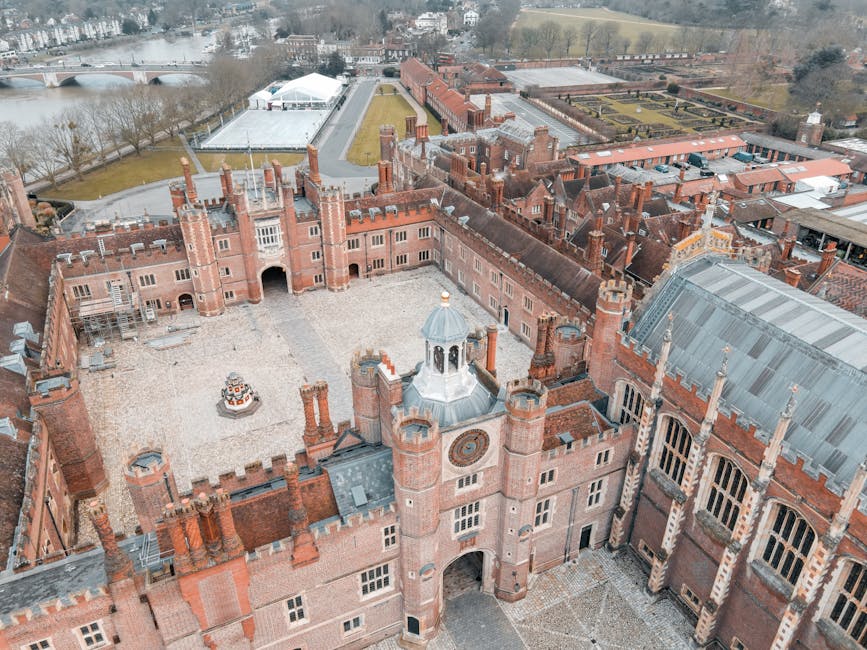 Aerial view of a historic brick castle with multiple towers, battlements, and ornate architectural details. The courtyard within the castle walls features a gravel surface and a small decorative fount