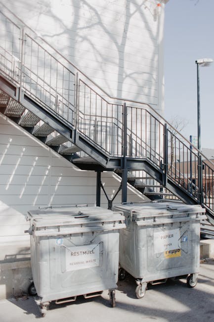Outside a building, two large grey wheeled trash bins marked as 'RECYCLING' and 'GENERAL WASTE' are positioned on a paved area near a metal staircase with an orange-brown railing. The staircase leads up to an upper level, casting shadows on the white exterior wall of the building. The scene appears to be part of a home relocation or moving process managed by Man and Van Hampton, with the bins possibly used to dispose of packing materials or waste generated during packing and loading activities. The area is well-lit by natural sunlight, with minimal other objects present, emphasizing the practical aspects of moving logistics and waste disposal in a house removal context.