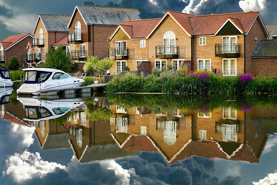 A row of modern, multi-storey brick residential buildings with pitched roofs and small balconies in front, situated along a canal or river. The foreground shows a boat docked at the water's edge, with its reflection visible on the calm, mirror-like surface of the water. Surrounding the scene are lush green trees and flowering plants, with some purple flowers near the water. The sky above is partly cloudy, casting natural light onto the buildings and water reflections. The setting indicates a peaceful riverside location suitable for house removals and home relocations, as managed by Man and Van Hampton, with the preparation and transport of household furniture and boxes around the property for moving day.