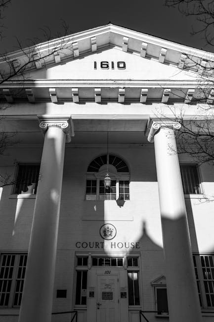 A black and white photograph of a traditional courthouse building featuring a prominent pediment with the number '1610' displayed at the top. The structure is supported by two large, classical columns with ornate capitals, framing the entrance below. A hanging lantern light is centered above the doorway, casting shadows on the façade. The front door is marked with the words 'COURT HOUSE' and the number '101' near the entrance, with small windows on either side. The building is set against a background of leafless trees and an overcast sky, emphasizing the historic architecture. This image exemplifies a formal judicial or government building, with clear details suitable for a professional description related to property, legal, or civic services, such as removals or house relocations executed by companies like Man and Van Hampton, which may involve access to such properties.