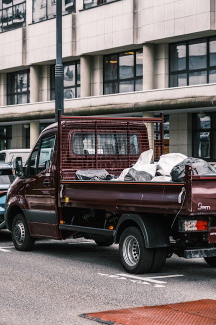 A dark red flatbed van parked on the street outside a modern commercial building with large curved windows. The vehicle's cargo area is filled with various household belongings, including cardboard boxes, plastic-wrapped items, and packaged furniture, all arranged for home relocation. Some items are covered with protective blankets, and the load extends to the edge of the van’s sides. The van features a metal mesh partition behind the cab to secure the load during transit. The surrounding environment includes other parked vehicles and a streetlamp, with the scene captured during daylight hours. This image illustrates the process of furniture transport and packing involved in removals services, as offered by Man and Van Hampton, which is relevant to understanding the typical items and methods used during home removals, especially when addressing hidden costs in removal quotes. This visual highlights the loading process and collection of belongings for a house move in Hampton.