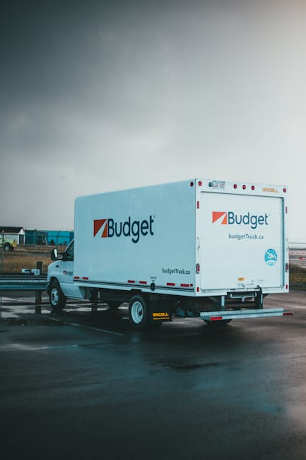 A white Budget moving truck parked on a wet asphalt surface, with its rear doors closed and the side panel displaying the Budget logo and website. The vehicle is positioned near the entrance of a property in a residential or commercial area, ready for house removals or furniture transport. In the background, there are other trucks and industrial buildings under a cloudy sky, indicating a loading or unloading process as part of a home relocation or packing and moving service. The area appears to be a designated loading zone, with some elements such as barriers or pavement markings visible. The scene captures the practical aspects of professional removals, including vehicle loading and transportation, essential for careful furniture transport during house moves managed by companies like Man and Van Hampton. The overall setting emphasizes efficient moving logistics within the context of understanding removal quote costs, as highlighted in the page about hidden costs in Hampton removal quotes.