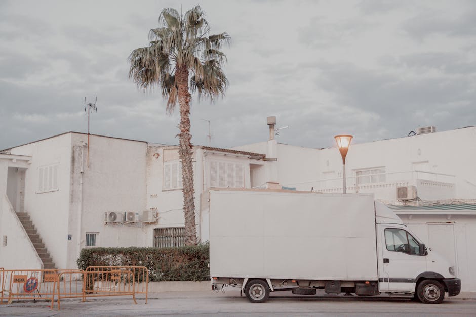 A dark red flatbed van parked on the street outside a modern commercial building with large curved windows. The vehicle's cargo area is filled with various household belongings, including cardboard boxes, plastic-wrapped items, and packaged furniture, all arranged for home relocation. Some items are covered with protective blankets, and the load extends to the edge of the van’s sides. The van features a metal mesh partition behind the cab to secure the load during transit. The surrounding environment includes other parked vehicles and a streetlamp, with the scene captured during daylight hours. This image illustrates the process of furniture transport and packing involved in removals services, as offered by Man and Van Hampton, which is relevant to understanding the typical items and methods used during home removals, especially when addressing hidden costs in removal quotes. This visual highlights the loading process and collection of belongings for a house move in Hampton.