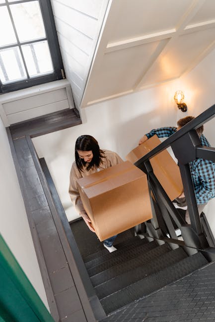 A woman with dark hair, wearing a beige sweater, is walking up a narrow staircase carrying a large cardboard moving box. Behind her, another person in a blue checkered shirt is partially visible, also holding a box. The staircase is situated inside a residential property with white walls, and natural light is coming through a large window at the top of the stairway. The stair treads are black with a textured surface, and a black handrail runs along the right side of the staircase. The scene captures the process of home relocation, where packing and furniture transport are underway, reflecting the challenges of navigating narrow stairs during house removals. The inside environment is well-lit, emphasizing the careful handling of household items in the moving process, with cardboard boxes and protective packaging materials visible, supported by professional removals services such as Man and Van Hampton.