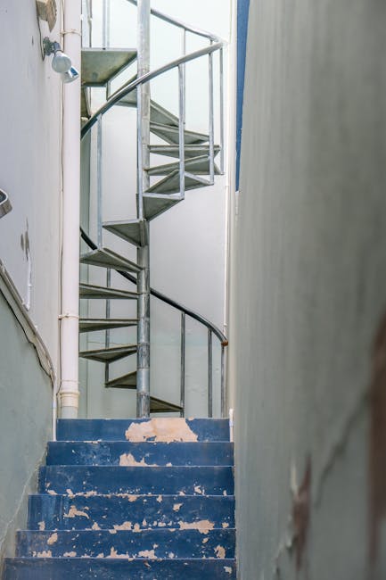 A narrow, steep staircase inside a property with worn blue-painted concrete steps, showing chipped paint and signs of use. The stairway is enclosed by white and light grey walls, with a simple metal handrail along the side. At the top of the stairs, a metal spiral staircase with open risers and a thin vertical pole in the centre leads upward, indicating a multi-level layout. Adjacent to the staircase, a white PVC pipe runs vertically along the wall, and a doorframe is partially visible on the left. The environment appears aged and in need of refurbishment, typical of older houses requiring careful handling during a home relocation process. This image exemplifies common challenges faced during furniture transport and packing in properties with narrow staircases, where professional removals like those offered by Man and Van Hampton assist with safe and efficient loading and moving operations in such constrained spaces.