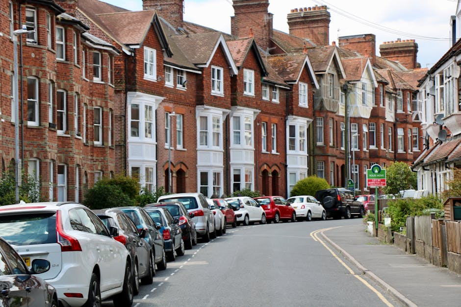 A suburban residential street with a row of red-brick Victorian-style terraced houses featuring bay windows and pitched roofs. Parked along the curb are various modern cars in different models and colours, including white, silver, red, and black. The road is narrow and slightly curved, with double yellow lines marking the side of the street. On the right, a green parking permit sign displays the words 'Your Home' alongside a circular logo, indicating parking restrictions or permissions related to local council regulations. The area is well-lit with natural daylight, and some greenery, including bushes and small trees, can be seen in front of the houses and along the pavement. This scene captures a typical home relocation or furniture transport setting during the daytime, with no visible activity but emphasizing a quiet, residential environment suitable for house removals and moving services, as provided by Man and Van Hampton.
