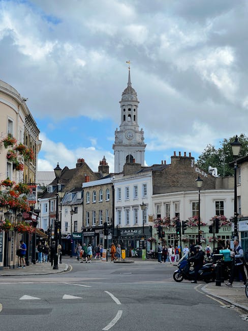 A busy street scene in Hampton featuring a historic clock tower with a spire and weather vane in the background under a partly cloudy sky. The street is lined with multi-story buildings displaying a mix of architectural styles, with shopfronts on the ground level and residential or office windows above. Retail shops and cafes with signage and flower baskets hanging from lampposts are visible, along with pedestrians walking on the sidewalk. In the foreground, a vehicle, possibly an electric scooter or moped, is parked on the street near the curb. The area is illuminated by traditional black street lamps, and the overall scene depicts a lively, urban environment that could be involved in home relocation or furniture transport activities, consistent with house removals and moving services by Man and Van Hampton.