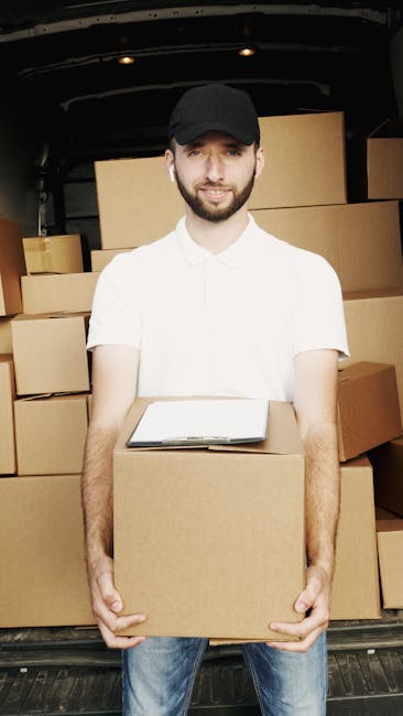 A man with a beard, wearing a black cap, white T-shirt, and blue jeans, is standing inside the open back of a van used for house removals. He is holding a cardboard box in front of him with both hands, and a clipboard rests on top of the box. Behind him, multiple packed cardboard boxes are stacked, some wrapped in plastic or placed in protective packaging. The van's interior appears to be spacious and is illuminated, showing the boxes arranged for loading or unloading. The image captures the process of furniture transport and packing during a home relocation, reflecting professional removals services by Man and Van Hampton. The setting suggests the completion of a packing or loading phase of a same-day moving process in Hampton, illustrating efficient logistics involved in furniture transport and house removals.