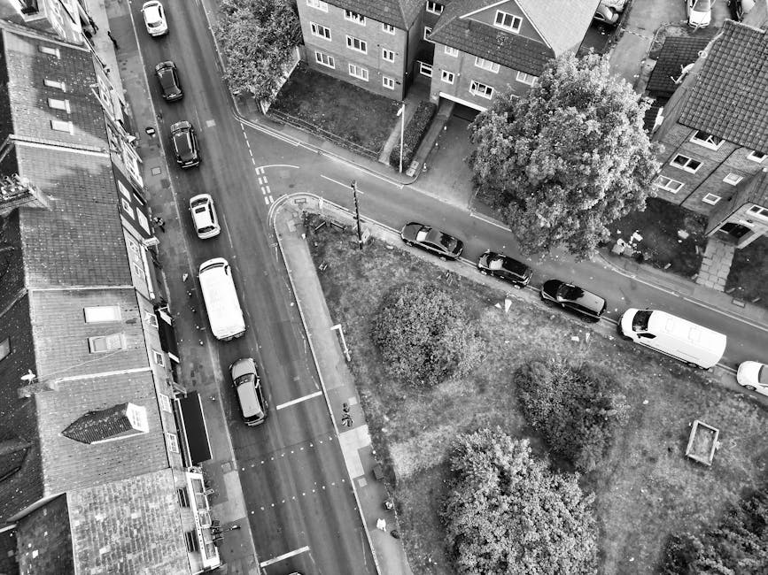 An aerial black-and-white photograph showing a residential street with parked cars along the curb and a small green park area with trees in the middle. On the right side, a man from Man and Van Hampton is seen in the process of loading or unloading moving boxes wrapped in cardboard and plastic from the back of a white moving van. The van is parked on the pavement near the park, with the rear doors open, revealing packed boxes and perhaps furniture wrapped in moving blankets. The surrounding scene features multi-storey brick houses with pitched roofs, some with visible windows and doorways. The street is relatively quiet with a few additional vehicles parked along the road, and the environment suggests an organized home relocation or furniture transport operation as part of a house removals service.