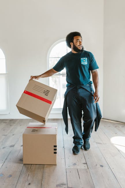 A man with dark curly hair and a beard, wearing a navy blue T-shirt with a company logo on the chest and dark trousers, is inside a well-lit room with white walls and wooden flooring. He is holding a cardboard box with red packing tape in his right hand, preparing to place it on top of another similar box positioned on the floor. The boxes are standard packing cartons, some with visible labels or barcodes, and they are arranged near a large arched window that allows natural light to illuminate the space. The man appears to be engaged in the home relocation process, involved in packing or furniture transport tasks as part of a professional moving service. The room is clear of other objects, with no furniture or appliances visible, emphasizing a stage of packing or loading during a house removal project managed by Man and Van Hampton, supporting house removals services in the Hampton area.