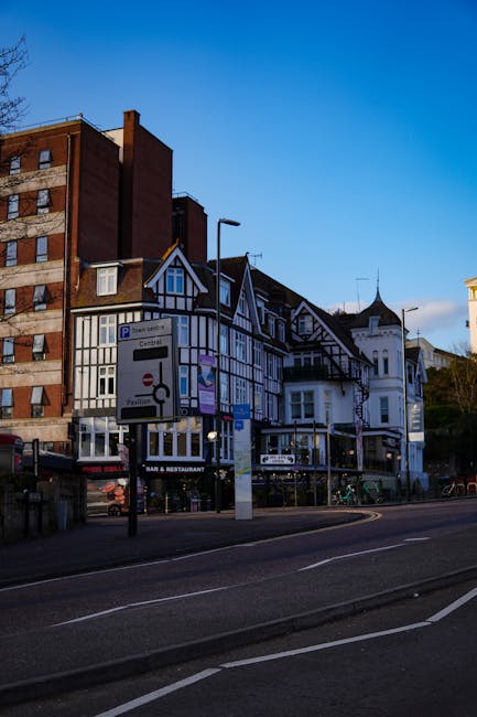 An aerial black-and-white photograph showing a residential street with parked cars along the curb and a small green park area with trees in the middle. On the right side, a man from Man and Van Hampton is seen in the process of loading or unloading moving boxes wrapped in cardboard and plastic from the back of a white moving van. The van is parked on the pavement near the park, with the rear doors open, revealing packed boxes and perhaps furniture wrapped in moving blankets. The surrounding scene features multi-storey brick houses with pitched roofs, some with visible windows and doorways. The street is relatively quiet with a few additional vehicles parked along the road, and the environment suggests an organized home relocation or furniture transport operation as part of a house removals service.